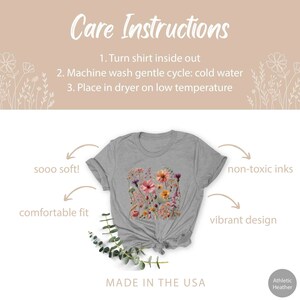 K&ouml;nnte beinhalten: Ein graues T-Shirt mit einem floralen Druckdesign. Das Shirt ist in den USA hergestellt und verwendet nicht-toxische Farben. Der Text "sooo soft!" und "comfortable fit" steht auf der linken Seite des Bildes. Der Text "non-toxic inks" und "vibrant design" steht auf der rechten Seite des Bildes. Der Text "MADE IN THE USA" steht am unteren Rand des Bildes.