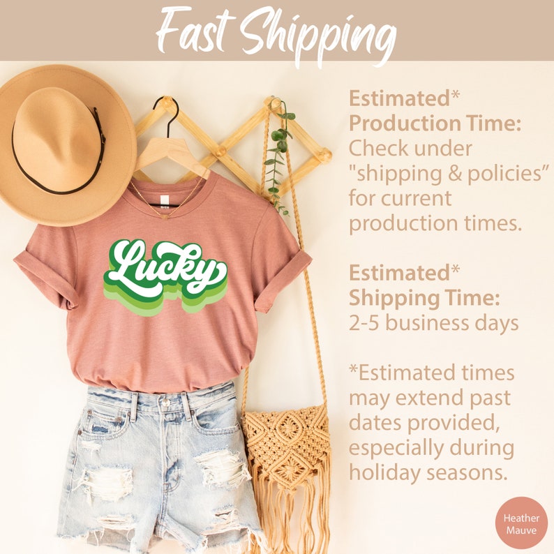 May include: A heather mauve t-shirt with a green retro-style graphic that says "Lucky". The shirt is hanging on a wooden rack with a straw hat and a woven bag.