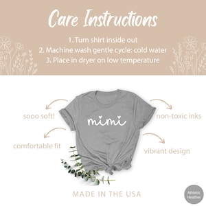 May include: Gray t-shirt with the word "mimi" in white script with two small hearts above the word. The shirt is shown on a white background with a green plant in the foreground. The text "sooo soft!" is written above the left shoulder of the shirt, "comfortable fit" is written below the left shoulder, "non-toxic inks" is written above the right shoulder, and "vibrant design" is written below the right shoulder. The text "MADE IN THE USA" is written below the shirt. The text "Athletic Heather" is written in the bottom right corner.