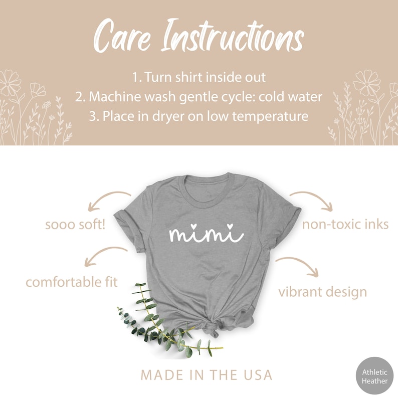 May include: Gray t-shirt with the word "mimi" in white script with two small hearts above the word. The shirt is shown on a white background with a green plant in the foreground. The text "sooo soft!" is written above the left shoulder of the shirt, "comfortable fit" is written below the left shoulder, "non-toxic inks" is written above the right shoulder, and "vibrant design" is written below the right shoulder. The text "MADE IN THE USA" is written below the shirt. The text "Athletic Heather" is written in the bottom right corner.