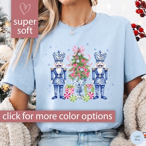 May include: Light blue t-shirt with a watercolor illustration of two nutcracker figurines flanking a Christmas tree with pink ornaments and a wreath. The text "super soft" is in a pink heart shape at the top left of the image.