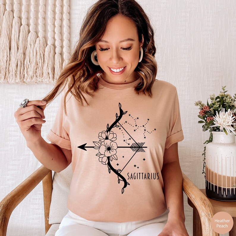 May include: A heather peach t-shirt with a black floral design featuring an arrow, bow, and the constellation Sagittarius. The text "SAGITTARIUS" is printed below the design.