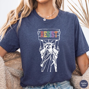 May include: A navy blue t-shirt with a white graphic of the Statue of Liberty holding a sign that says "RESIST" in rainbow colors.