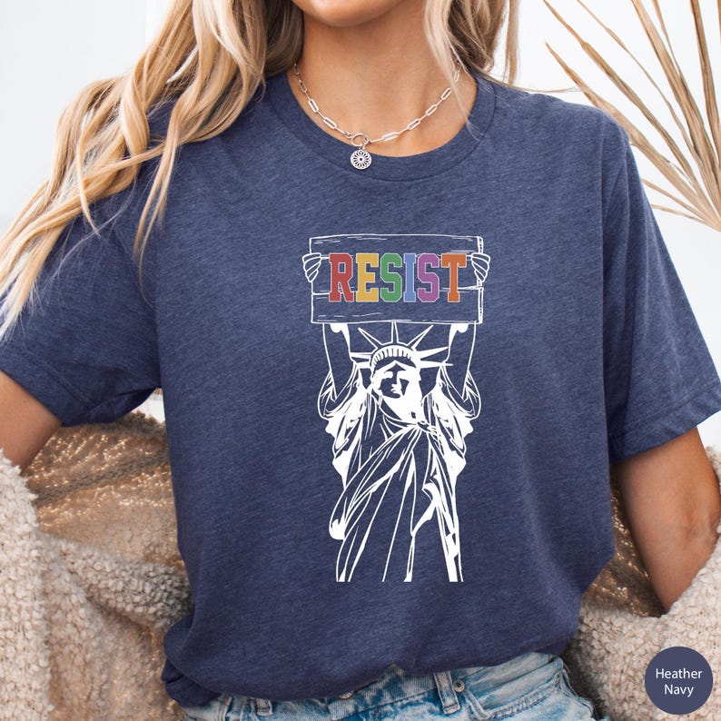 May include: A navy blue t-shirt with a white graphic of the Statue of Liberty holding a sign that says "RESIST" in rainbow colors.
