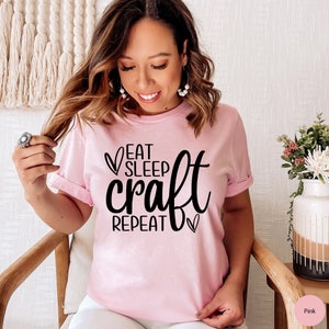 May include: A pink t-shirt with black text that reads "Eat Sleep Craft Repeat" with a heart symbol before and after the word "Craft".