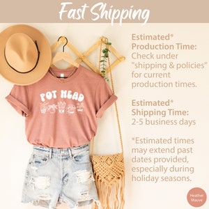 May include: A heather mauve t-shirt with the text "POT HEAD" in white lettering. The shirt features a graphic of potted plants. The shirt is hanging on a wooden rack with a straw hat and a woven bag.