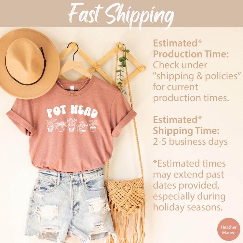 May include: A heather mauve t-shirt with the text "POT HEAD" in white lettering. The shirt features a graphic of potted plants. The shirt is hanging on a wooden rack with a straw hat and a woven bag.