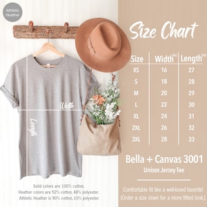 May include: A light gray Bella + Canvas 3001 Unisex Jersey Tee hanging on a wooden rack. The image includes a size chart with measurements in inches, a brown hat, and a beige purse with flowers. The shirt is 100% cotton.