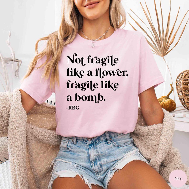 May include: A pink t-shirt with the text "Not fragile like a flower, fragile like a bomb. -RBG" printed in black.