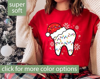 May include: Red t-shirt with a Christmas-themed graphic. The design features a tooth wearing a Santa hat and decorated with colorful Christmas lights. The shirt also has snowflakes and the text "click for more color options".