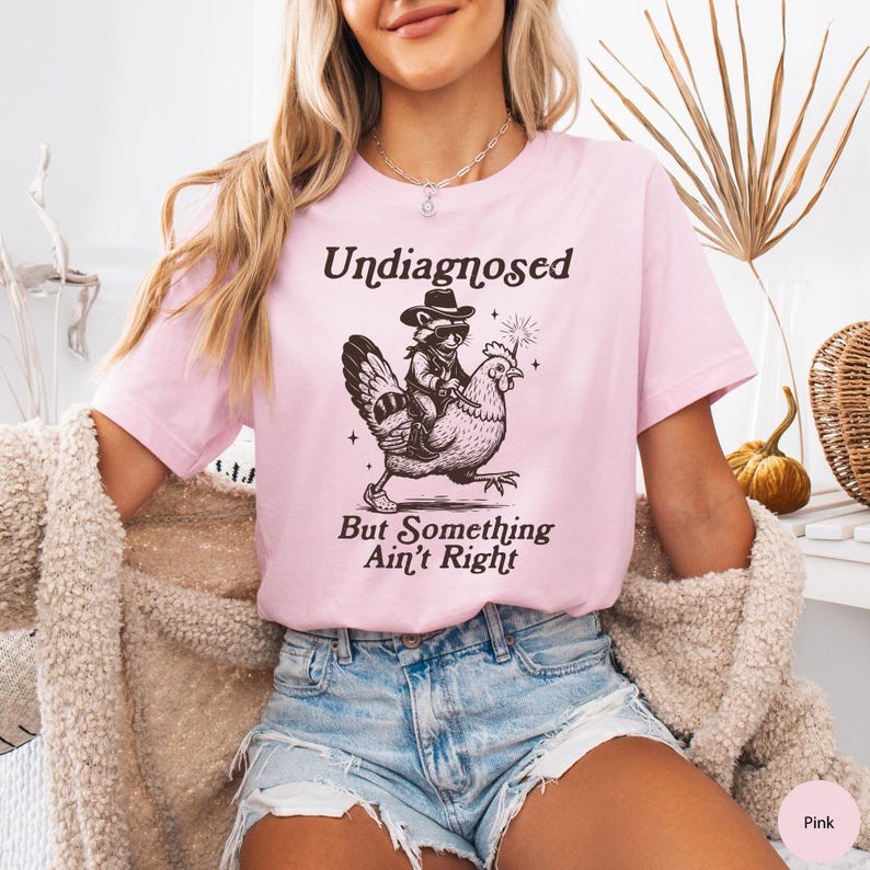May include: A light pink t-shirt with the text "Undiagnosed But Something Ain't Right" and a graphic of a raccoon riding a chicken. The raccoon wears sunglasses and a cowboy hat. The shirt is made of a soft material.