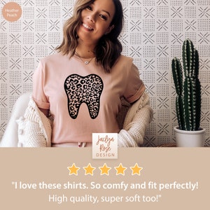 May include: A pink t-shirt with a black tooth graphic featuring a leopard print design. The shirt is worn by a person and the text "Jaclyn Rose DESIGN" and five stars are visible below the shirt. The text "I love these shirts. So comfy and fit perfectly! High quality, super soft too!" is below the stars.