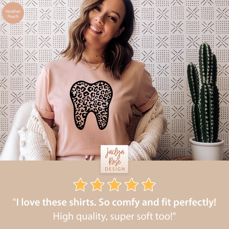 May include: A pink t-shirt with a black tooth graphic featuring a leopard print design. The shirt is worn by a person and the text "Jaclyn Rose DESIGN" and five stars are visible below the shirt. The text "I love these shirts. So comfy and fit perfectly! High quality, super soft too!" is below the stars.