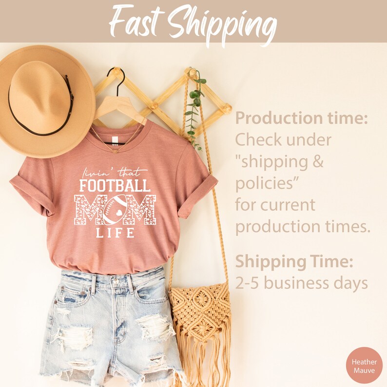 May include: A heather mauve t-shirt with a white graphic that says "Livin' that Football Mom Life" with a football and floral design. The shirt is hanging on a wooden hanger with a brown hat and a woven bag.