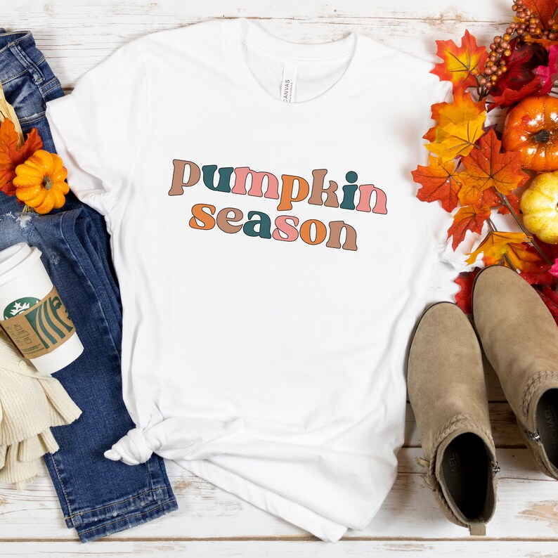 Puede incluir: Camiseta blanca con el texto "pumpkin season" en letras retro de colores. La camiseta est&aacute; colocada sobre una superficie de madera con un par de botines marrones y follaje de oto&ntilde;o.
