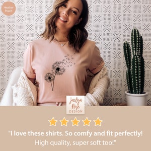 May include: A woman wearing a heather peach t-shirt with a dandelion graphic. The shirt has the text "Jaclyn Rose DESIGN" on the bottom. The woman is smiling and giving a thumbs up. The background is a white and black patterned wall.