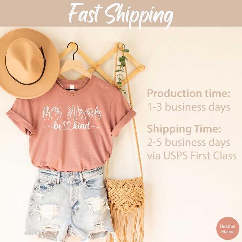 May include: A heather mauve t-shirt with a white graphic of the sign language for "be kind". The shirt is hanging on a wooden hanger with a straw hat and a woven bag.