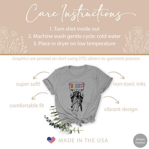 May include: A grey t-shirt with a graphic of the Statue of Liberty holding a sign that says "Resist" in rainbow colors. The text "Made in the USA" is printed on the bottom of the shirt.