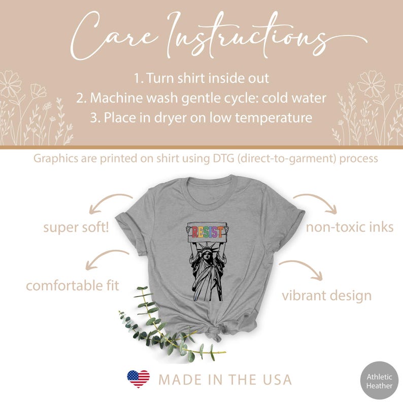 May include: A grey t-shirt with a graphic of the Statue of Liberty holding a sign that says "Resist" in rainbow colors. The text "Made in the USA" is printed on the bottom of the shirt.