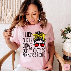 May include: Pink t-shirt with a graphic of a woman with a messy bun wearing sunglasses and a yellow crime scene tape headband. The text on the shirt reads "I like murder shows, comfy clothes and maybe 3 people."