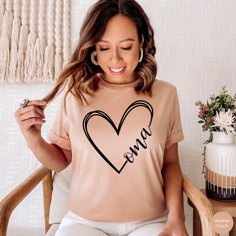 Pu&ograve; includere: Una maglietta rosa chiaro con un design a forma di cuore nero e la parola "oma" scritta all'interno del cuore.