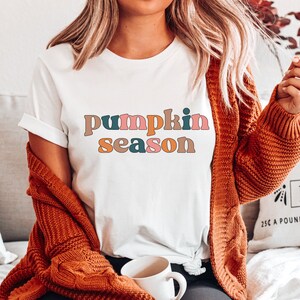 Puede incluir: Una camiseta blanca con el texto "pumpkin season" en letras coloridas de estilo retro. La camiseta la lleva puesta una persona con el pelo largo y rubio y un c&aacute;rdigan marr&oacute;n.