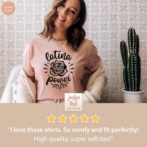 May include: A pink t-shirt with a black graphic design that says "latina power" with a rose graphic. The shirt is being worn by a person with a neutral expression. The background is a white wall with a black and white pattern.
