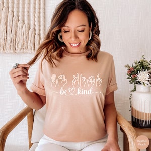 May include: A light pink t-shirt with a white graphic of the sign language for "be kind" with a heart in the middle of the word "be".