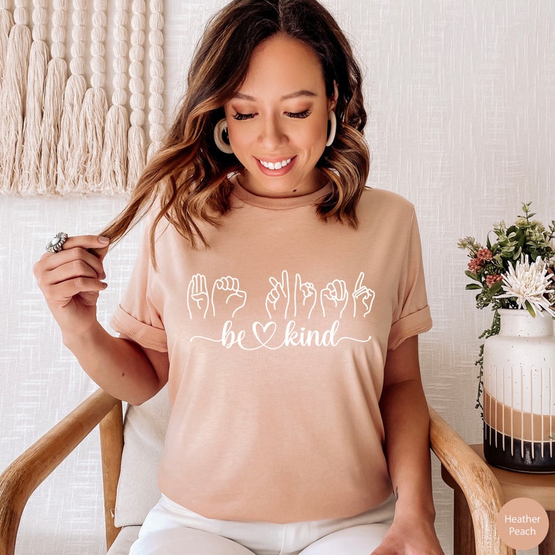 May include: A light pink t-shirt with a white graphic of the sign language for "be kind" with a heart in the middle of the word "be".