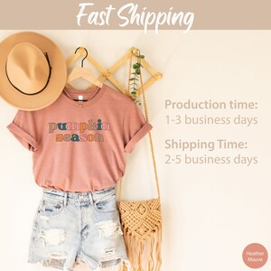 Puede incluir: Una camiseta de color malva con el texto "pumpkin season" en letras de bloque de colores. La camiseta est&aacute; colgada en un estante de madera con un sombrero marr&oacute;n y un bolso tejido.