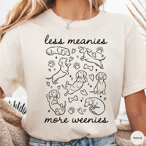 Op de afbeelding: Cr&egrave;mekleurig T-shirt met een zwarte afbeelding van teckels, pootafdrukken, harten en botten. De tekst "less meanies more weenies" staat in zwarte letters boven en onder de teckel illustraties.