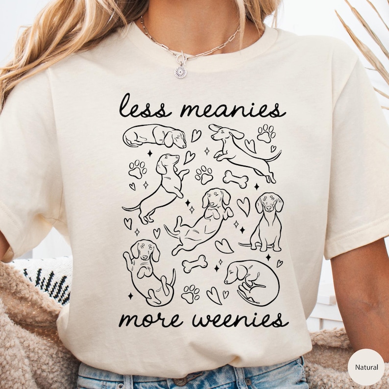 Op de afbeelding: Cr&egrave;mekleurig T-shirt met een zwarte afbeelding van teckels, pootafdrukken, harten en botten. De tekst "less meanies more weenies" staat in zwarte letters boven en onder de teckel illustraties.