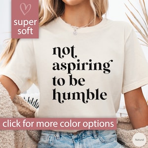 May include: A white t-shirt with the text "not aspiring to be humble" printed in black. The shirt is being worn by a person with long brown hair. The text "super soft" is printed in pink on the top left corner of the image.