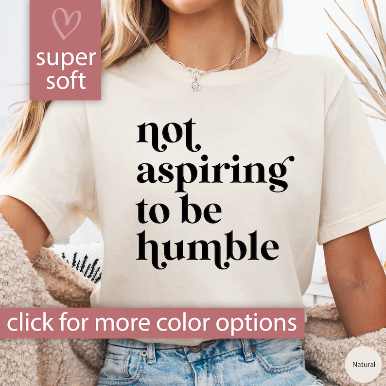 May include: A white t-shirt with the text "not aspiring to be humble" printed in black. The shirt is being worn by a person with long brown hair. The text "super soft" is printed in pink on the top left corner of the image.