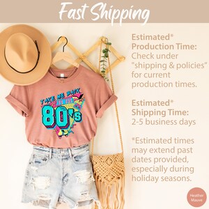 Pu&ograve; includere: Una t-shirt color malva con una stampa grafica retr&ograve; degli anni '80 che dice "Take Me Back to the 80s". La t-shirt &egrave; appesa a un appendiabiti di legno con un cappello di paglia e una borsa intrecciata.