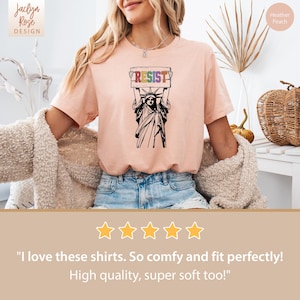 May include: A pink t-shirt with a graphic of the Statue of Liberty holding a sign that says "RESIST" in rainbow colors. The sign is held above her head. The t-shirt is a heather peach color.