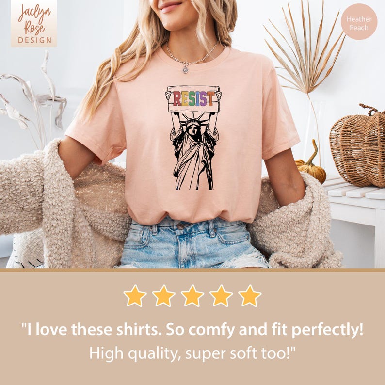 May include: A pink t-shirt with a graphic of the Statue of Liberty holding a sign that says "RESIST" in rainbow colors. The sign is held above her head. The t-shirt is a heather peach color.