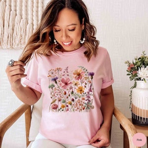 K&ouml;nnte beinhalten: Ein pinkfarbenes T-Shirt mit einem Blumendruck, der verschiedene Blumen in Rosa-, Gelb- und Lilat&ouml;nen zeigt. Die Blumen sind in einer Gruppe angeordnet und scheinen gepresst oder getrocknet zu sein.