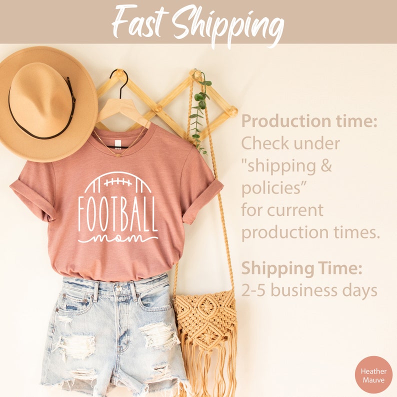 May include: A heather mauve t-shirt with a white graphic of a football and the text "FOOTBALL mom". The shirt is hanging on a wooden hanger with a straw hat and a woven bag.