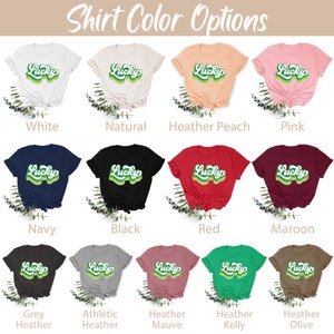 May include: A chart showing different t-shirt colors with the word "Lucky" in a retro green font on each shirt. The colors include white, natural, heather peach, pink, navy, black, red, maroon, grey heather, athletic heather, heather mauve, heather kelly, and heather olive.