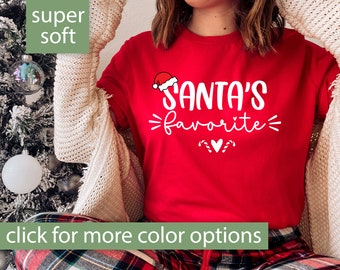 Könnte beinhalten: Rotes T-Shirt mit weißem Text, der "Santa's favorite" lautet, mit einer kleinen Santa-Mütze-Grafik über dem Text. Das Shirt wird von einer Person mit einem weißen Cardigan und einer rot-schwarz karierten Hose getragen.