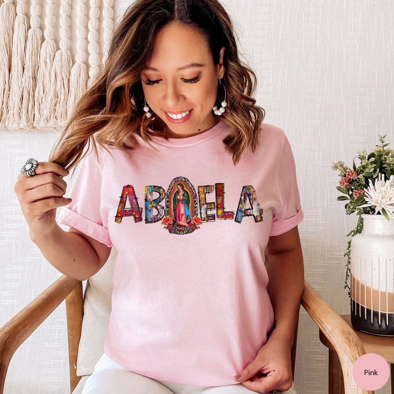 May include: Pink t-shirt with the word "ABUELA" in colorful letters with a religious image in the center.