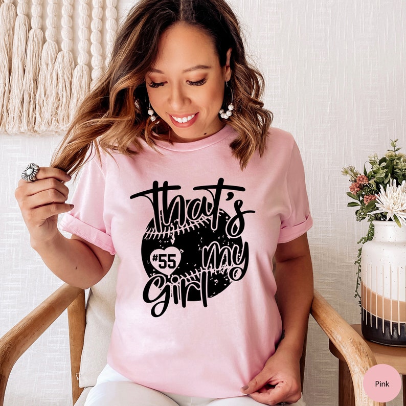 May include: Pink t-shirt with a black graphic of a baseball with a heart and the text "That's my girl #55".