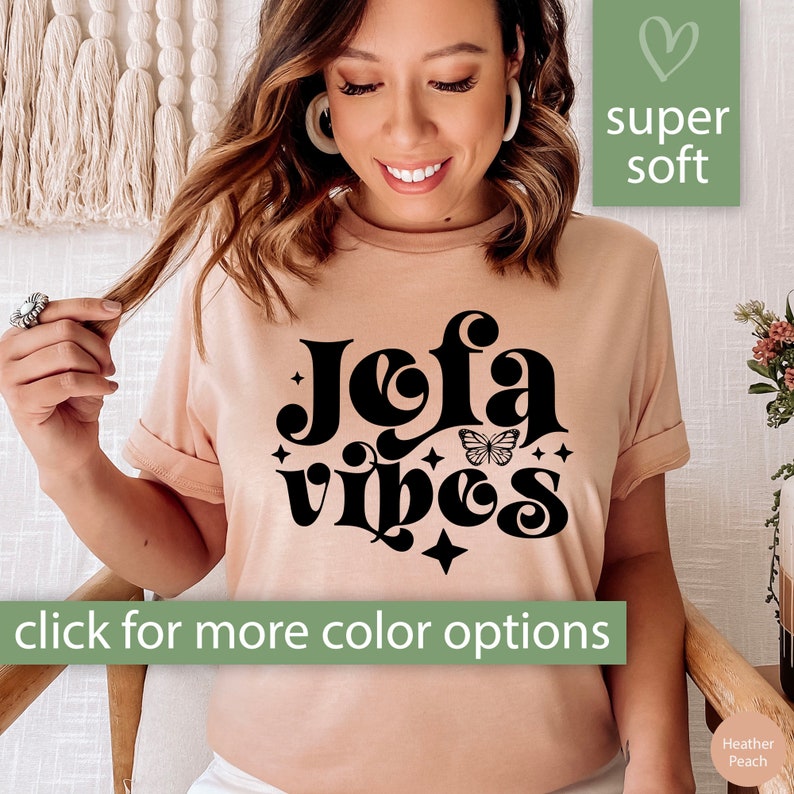 May include: Heather peach-colored t-shirt with the words "Jofa Vibes" in a black, retro font, a butterfly graphic, and stars. The shirt has rolled-up sleeves and the text "click for more color options". A green square with a heart and the words "super soft" is in the upper right.