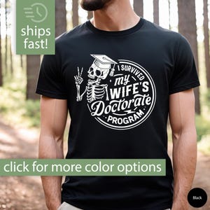 May include: Black t-shirt with a white graphic of a skeleton wearing a graduation cap and holding a peace sign. The graphic says "I Survived My Wife's Doctorate Program".