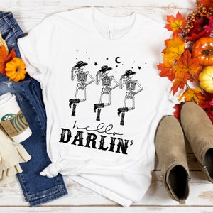 May include: White t-shirt with a graphic of three skeletons wearing cowboy hats and holding hammers. The text "hello darlin'" is printed below the skeletons.