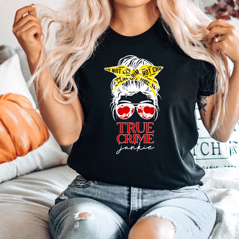 May include: Black t-shirt with a graphic of a woman with a messy bun wearing sunglasses and a yellow crime scene tape headband. The text "TRUE CRIME junkie" is printed below the graphic.