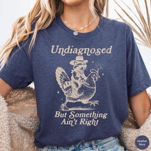 May include: Heather navy blue t-shirt with the text "Undiagnosed But Something Ain't Right." The shirt features a raccoon wearing a cowboy hat riding a chicken. The design is in a light tan color.