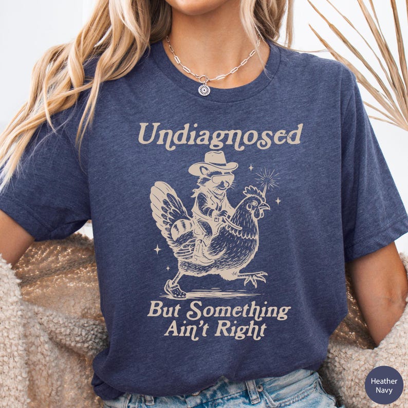 May include: Heather navy blue t-shirt with the text "Undiagnosed But Something Ain't Right." The shirt features a raccoon wearing a cowboy hat riding a chicken. The design is in a light tan color.