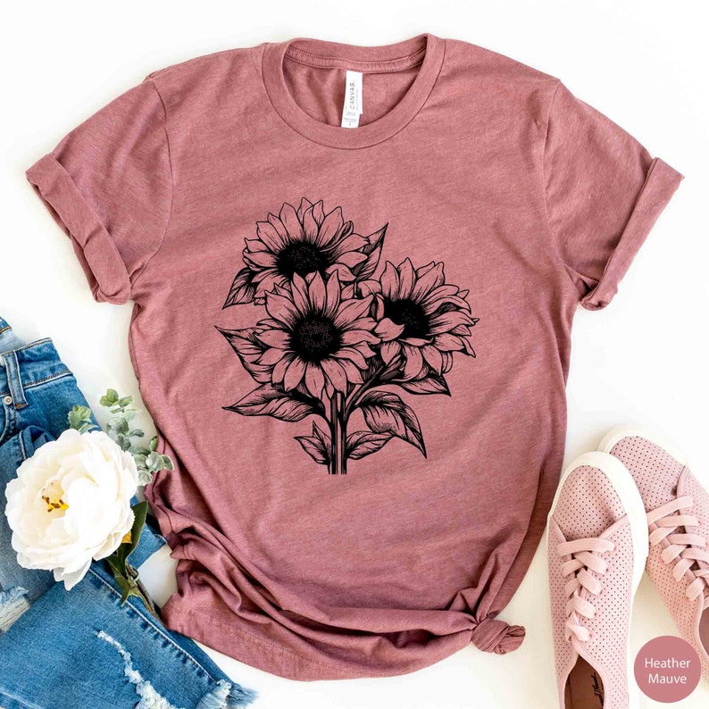 May include: Mauve-colored t-shirt featuring a black and white illustration of three sunflowers. The graphic design includes detailed petals and leaves. The shirt is made of soft material and has rolled-up sleeves.